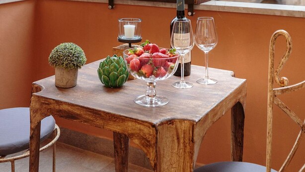 Une table en bois avec une bouteille, des verres, des fraises et une petite plante, le tout placé sur un balcon avec un mur de briques en arrière-plan.
