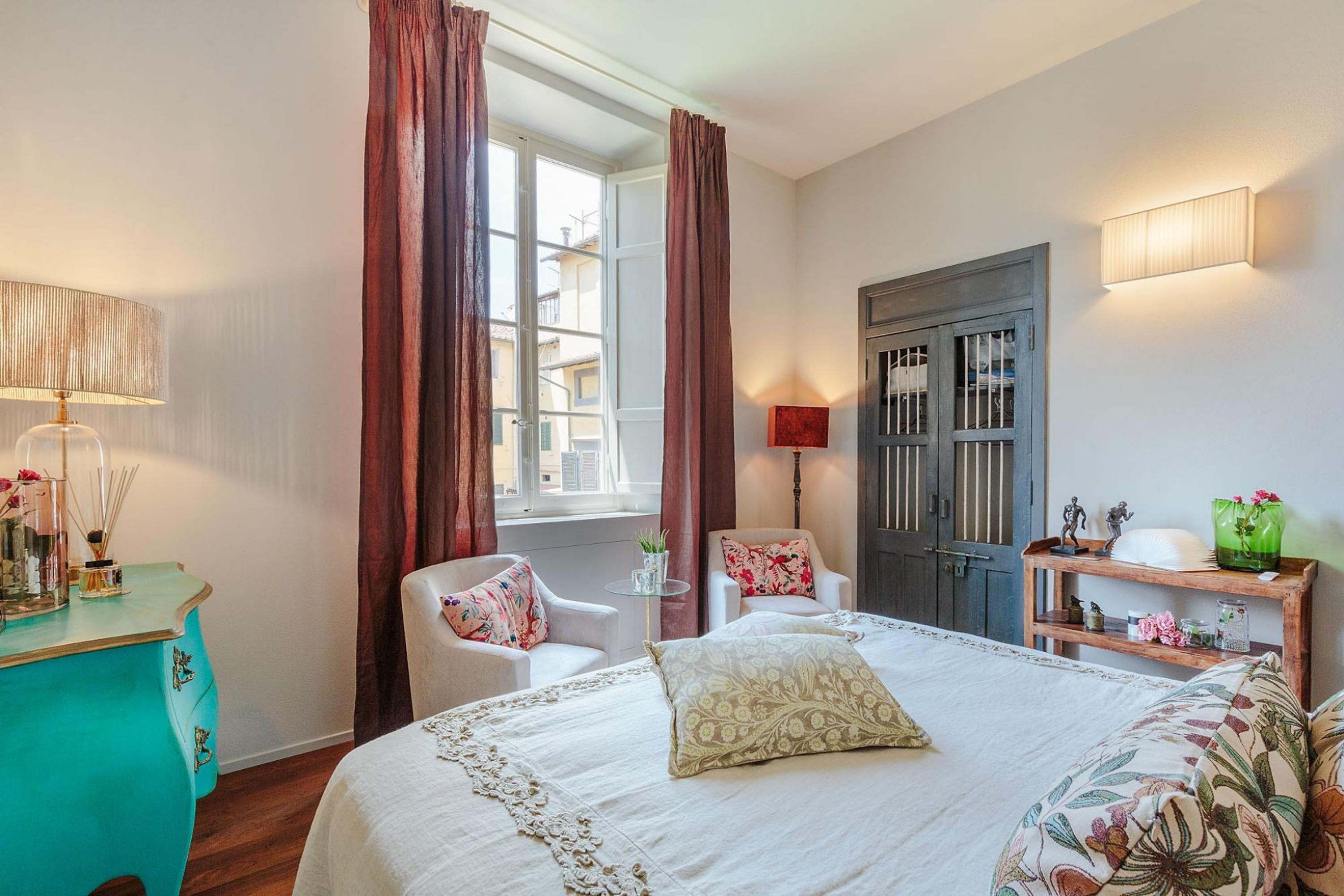 Une chambre à coucher comprenant un lit, deux chaises, un bureau, une fenêtre avec des rideaux, une porte et une lampe fixée au mur.