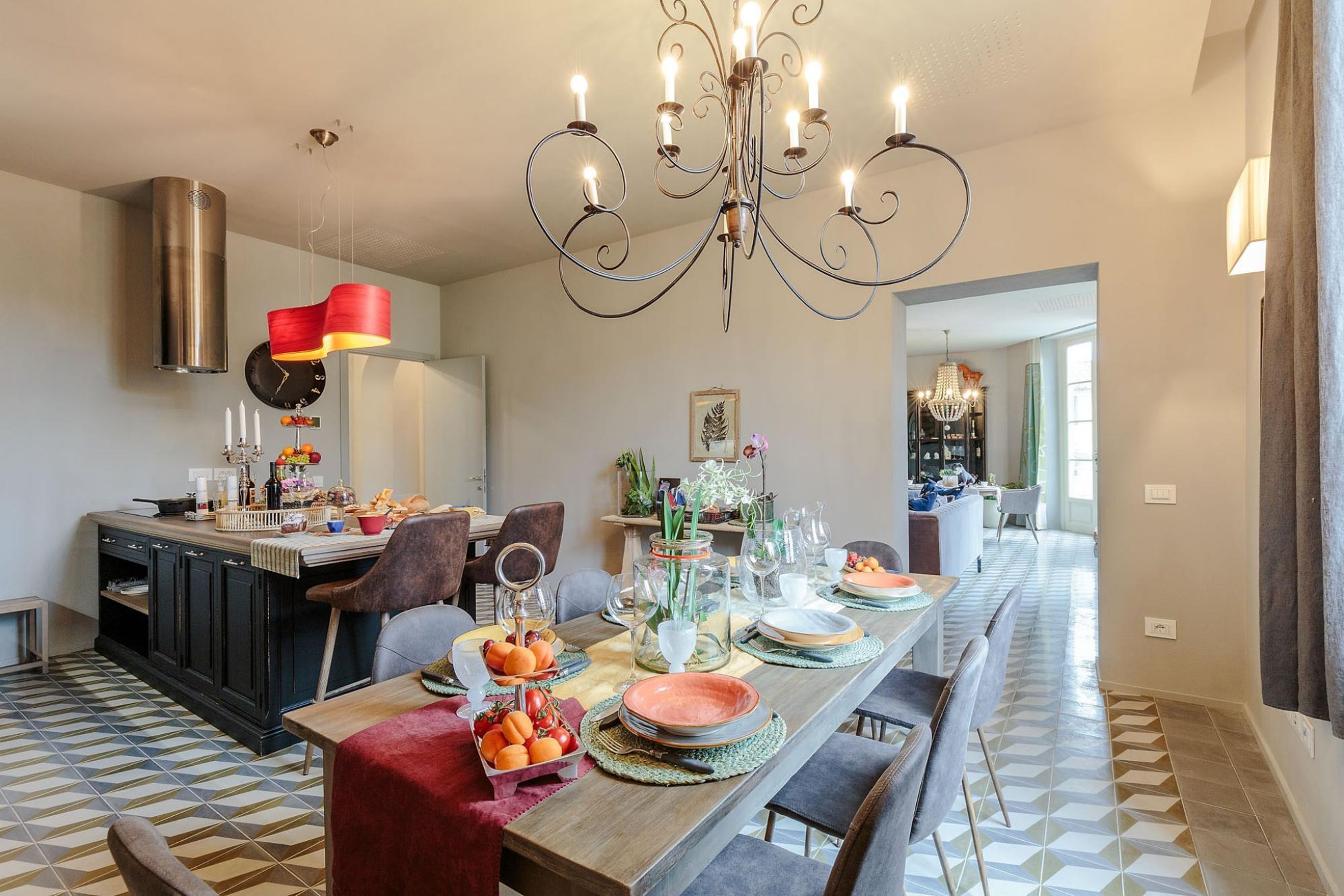 Comedor con una larga mesa dispuesta para una comida, un candelabro y una zona de cocina con estufa y armarios.