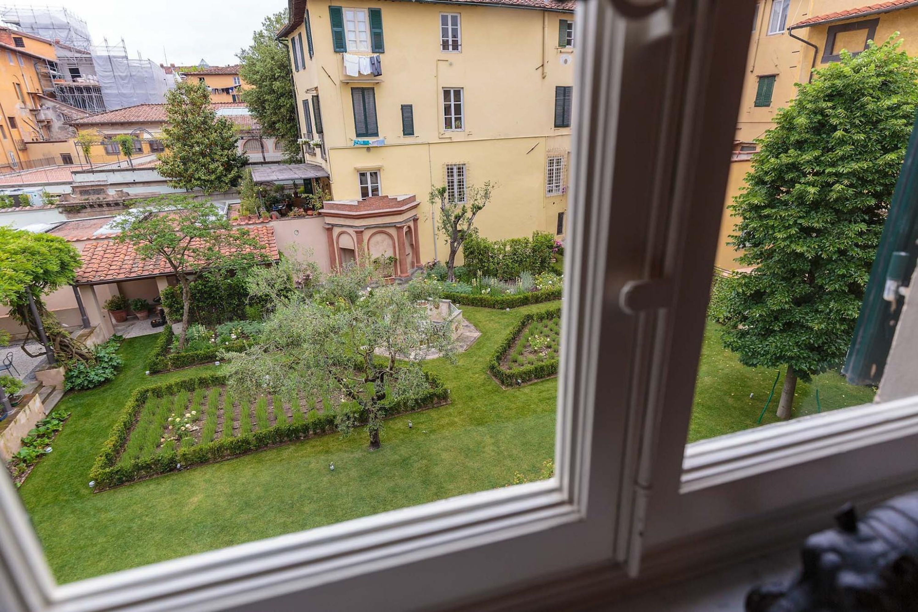 Un cortile con un giardino curato, una piccola fontana e un edificio con facciata gialla e persiane verdi.