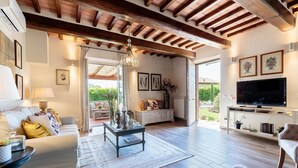 A spacious living room with a high ceiling, wooden beams, a large TV, and a view of the outdoors. - Villa Pemola in Capannori (Capannori)