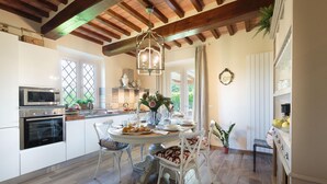 A modern kitchen with white cabinetry, a built-in oven, and a dining area with a round table and white chairs. - Villa Pemola in Capannori (Capannori)