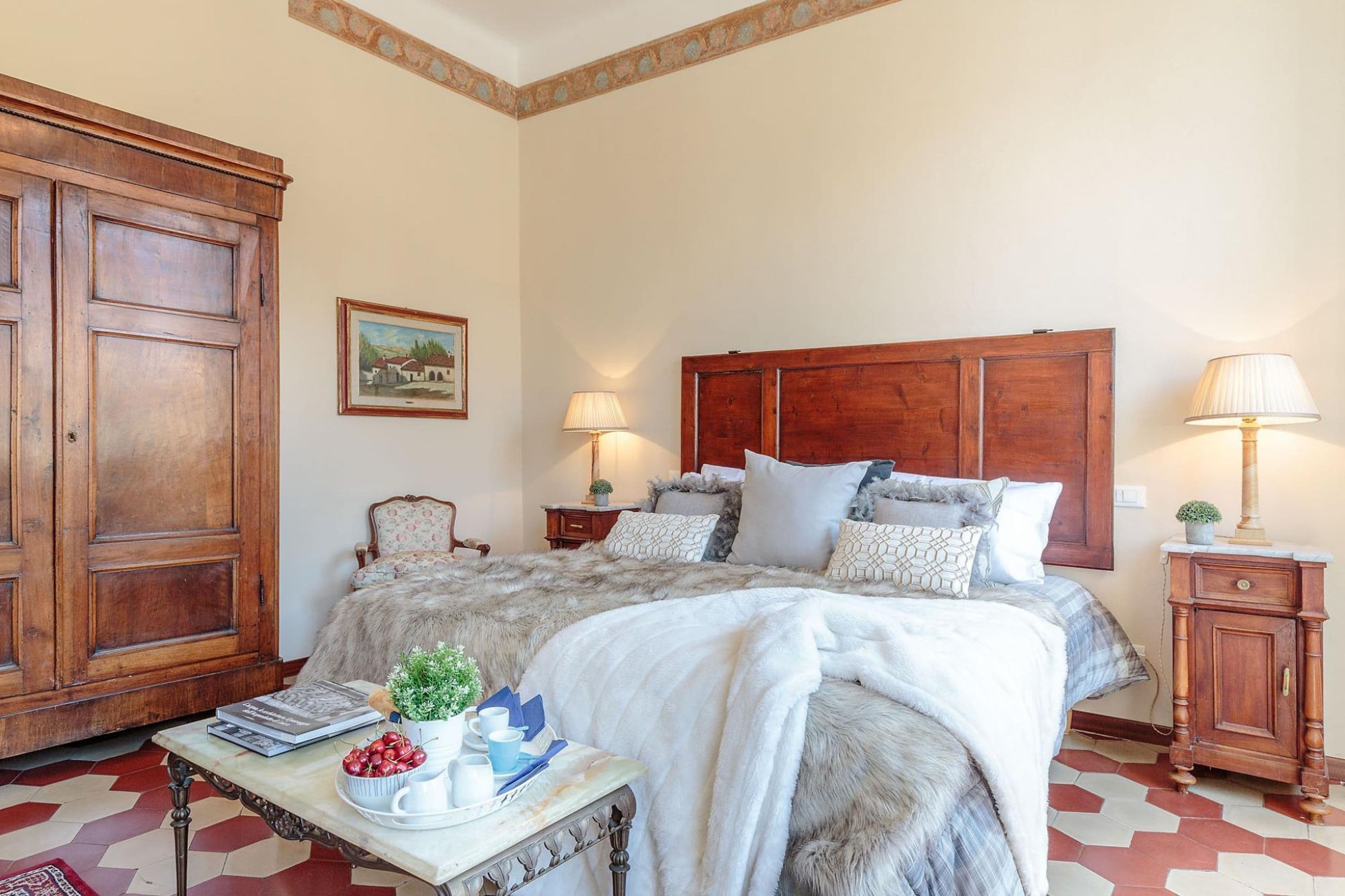 A bedroom with a large bed, wooden headboard, two bedside tables with lamps, a nightstand, a large wooden wardrobe, a framed picture on the wall, and a small table with a plant and books.