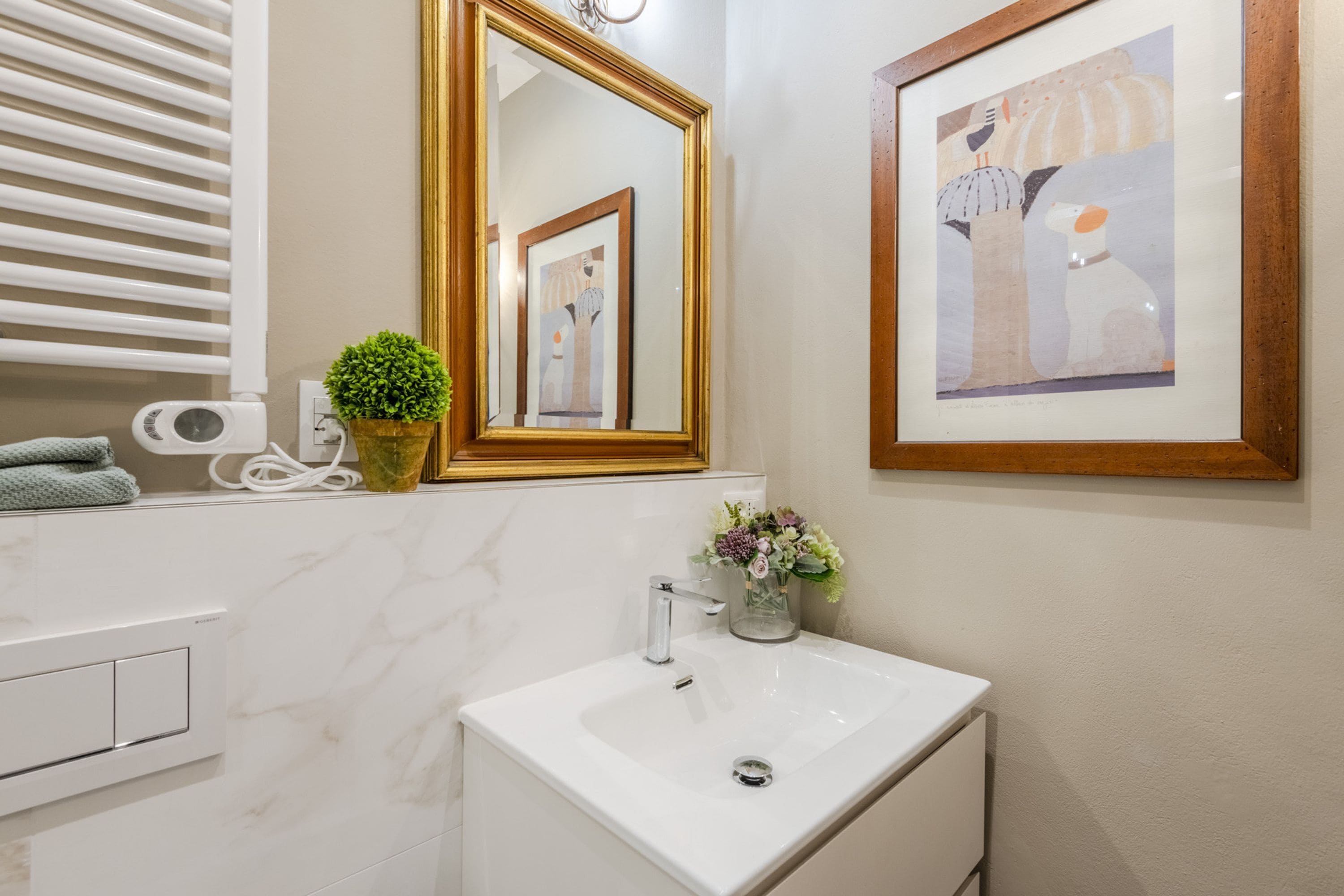 A bathroom with a white sink, a mirror with a gold frame, a framed artwork, a small potted plant, and a towel on the counter.