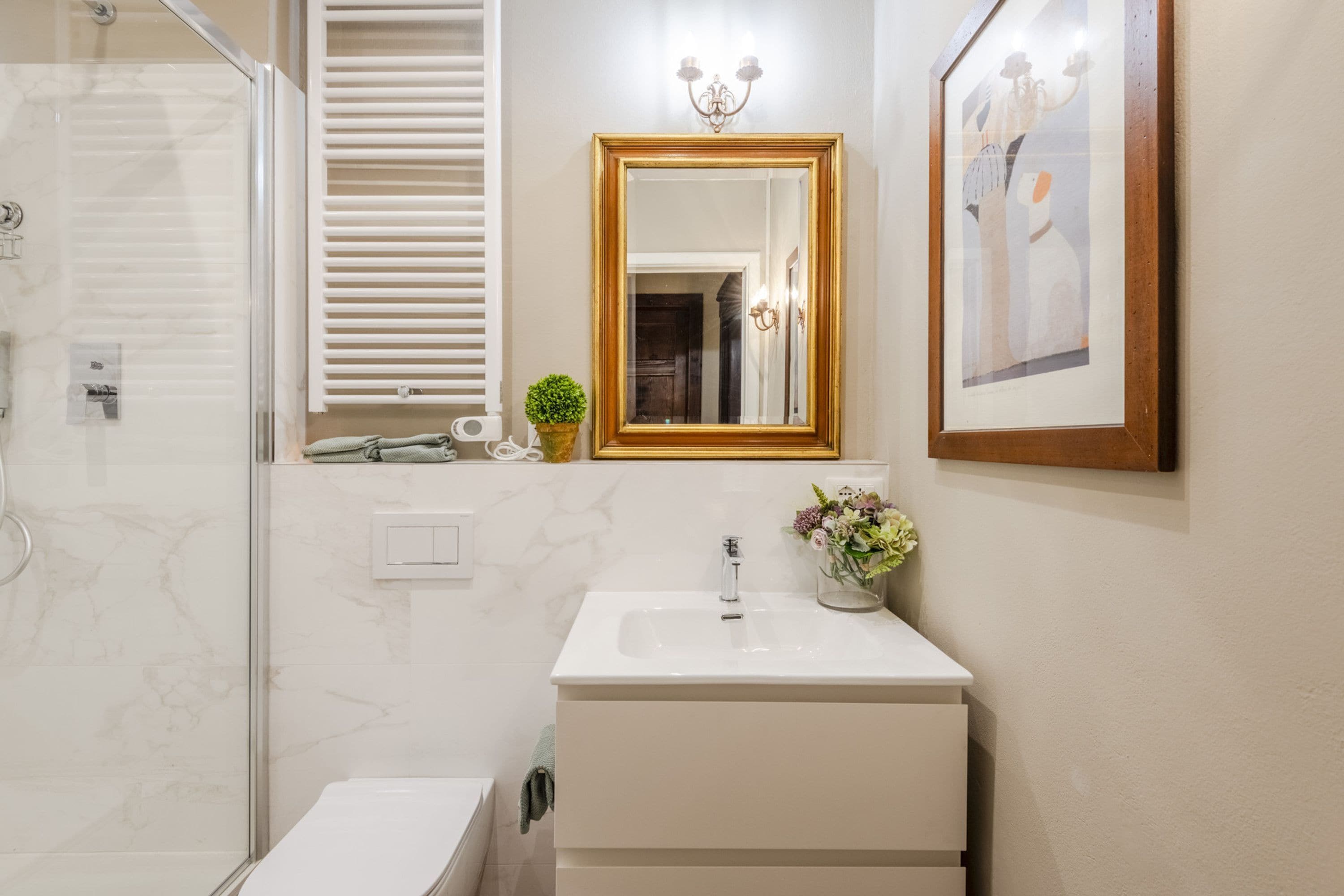A modern bathroom with a glass shower enclosure, a white sink with a vase of flowers, and a framed picture on the wall.