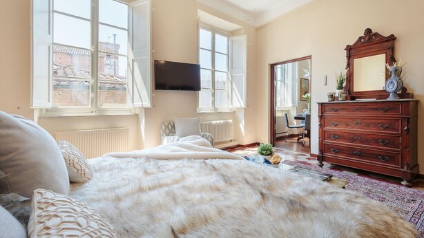 A bedroom with a large bed, a dresser, a TV mounted on the wall, and a window with a view of buildings. - Casa Santa Croce in Lucca (Lucca)