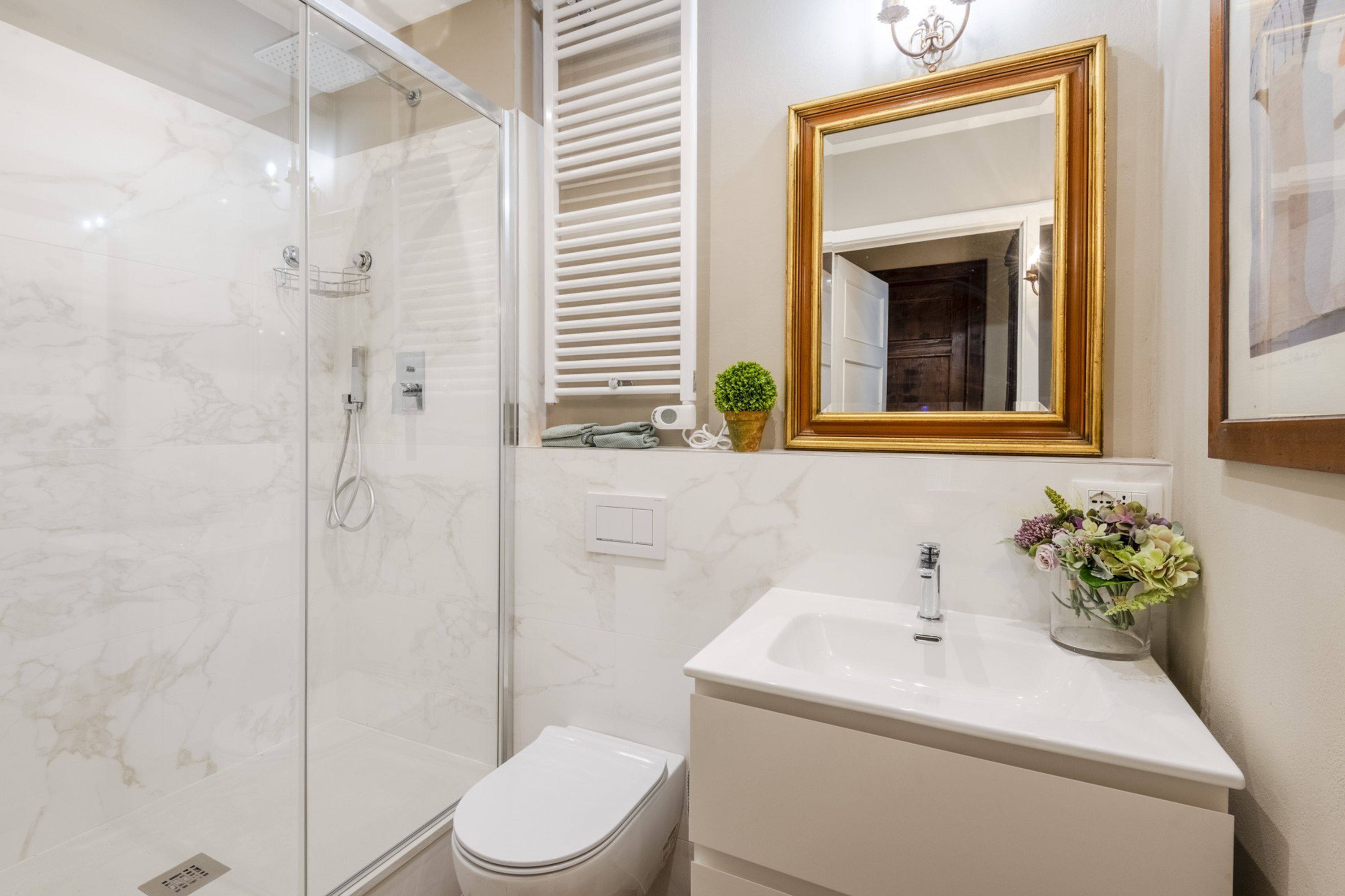 Ein modernes Badezimmer mit einer gläsernen Duschkabine, einer weißen Toilette, einem Waschbecken mit goldfarbenem Spiegelrahmen und einer Blumenvase.