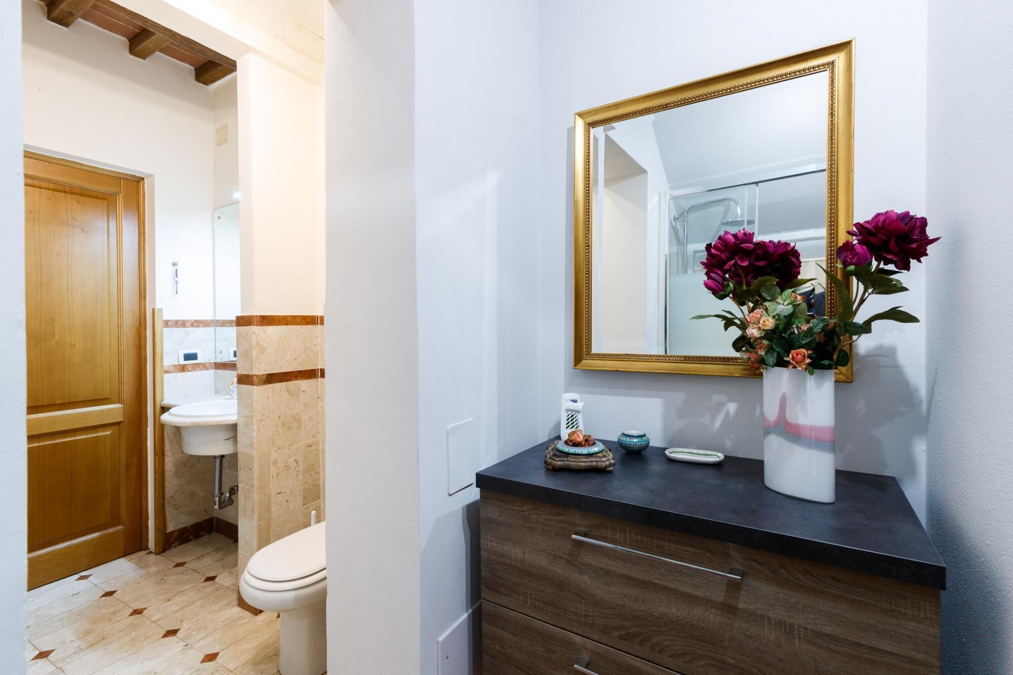 A bathroom with a wooden door, a sink, a toilet, and a vanity with a mirror and a vase of flowers.