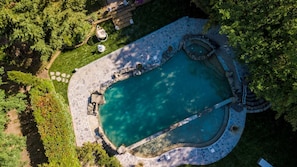 An aerial view of a swimming pool with a stone deck, surrounded by lush greenery and a small seating area.