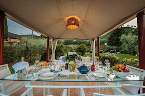An outdoor dining area with a glass table set for a meal, surrounded by greenery and a view of the countryside.
