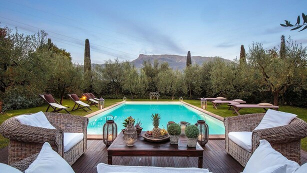 A pool area with wicker furniture and a table set with fruits and drinks, surrounded by greenery and overlooking a mountain. - Villa Label in Camaiore (Camaiore)