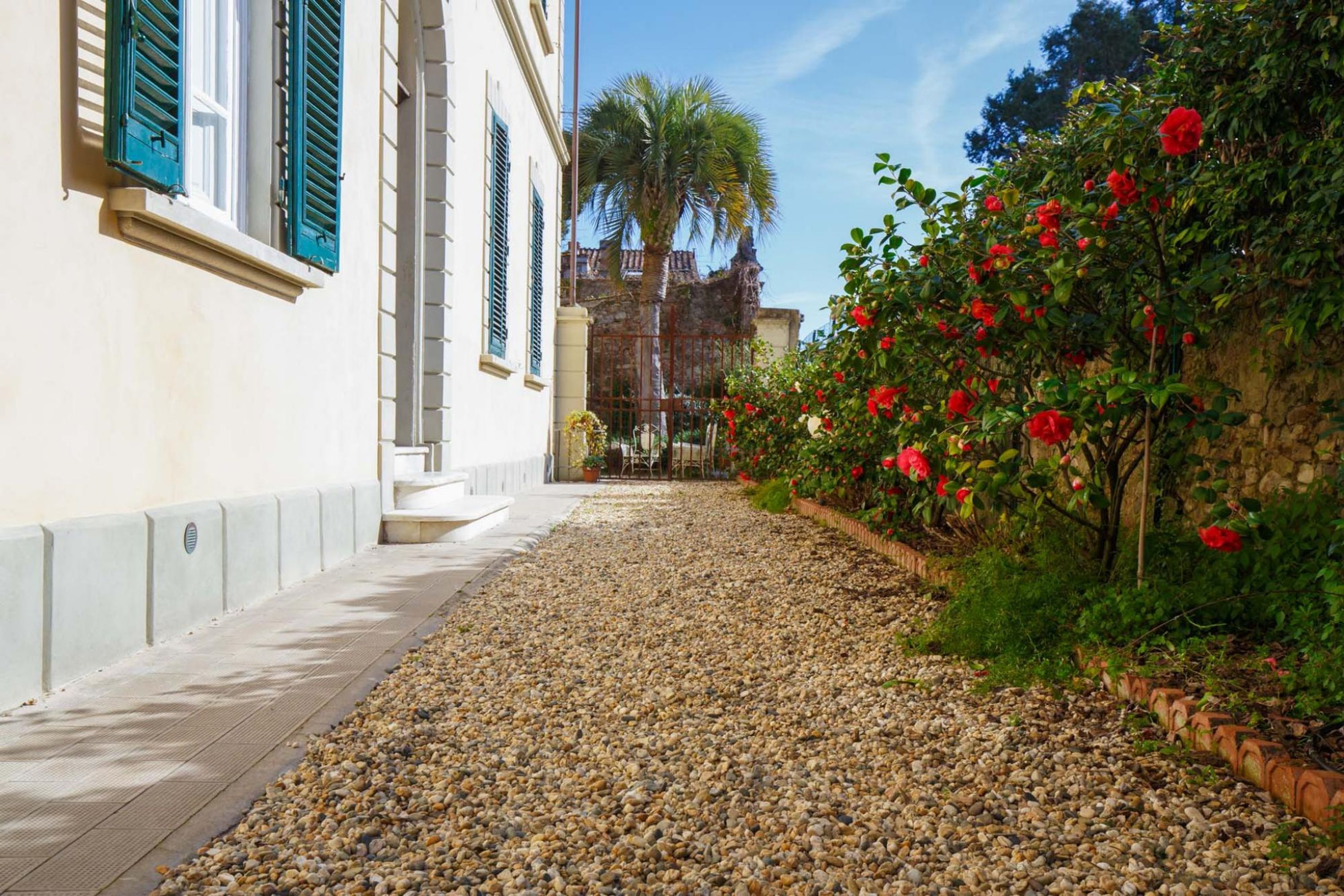 Un sendero de piedras que lleva a un edificio con contraventanas verdes y un jardín con flores rojas.