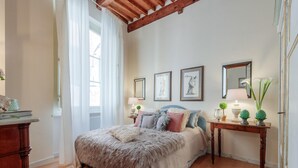 A bedroom with a large bed, a wooden dresser, a nightstand with a lamp, and a mirror on the wall. - Casa Ugolino in Lucca (Lucca)