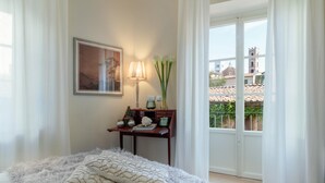 A bedroom with a large bed, a nightstand with a lamp, a framed picture, and a view of a cityscape through a glass door. - Casa Brumar in Lucca (Lucca)