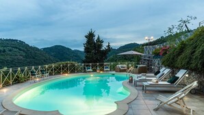 Une piscine entourée de chaises longues et d’un mur de pierre, le tout délimité par une clôture en métal et bordé d’arbres.