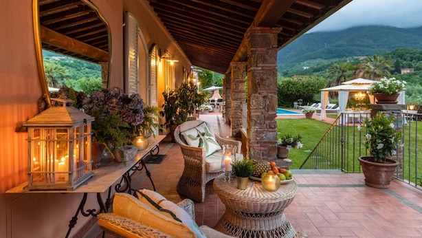 A covered outdoor seating area with wicker furniture, potted plants, and a view of a pool and mountains. - Villa Dana in Segromigno in Monte (Capannori)