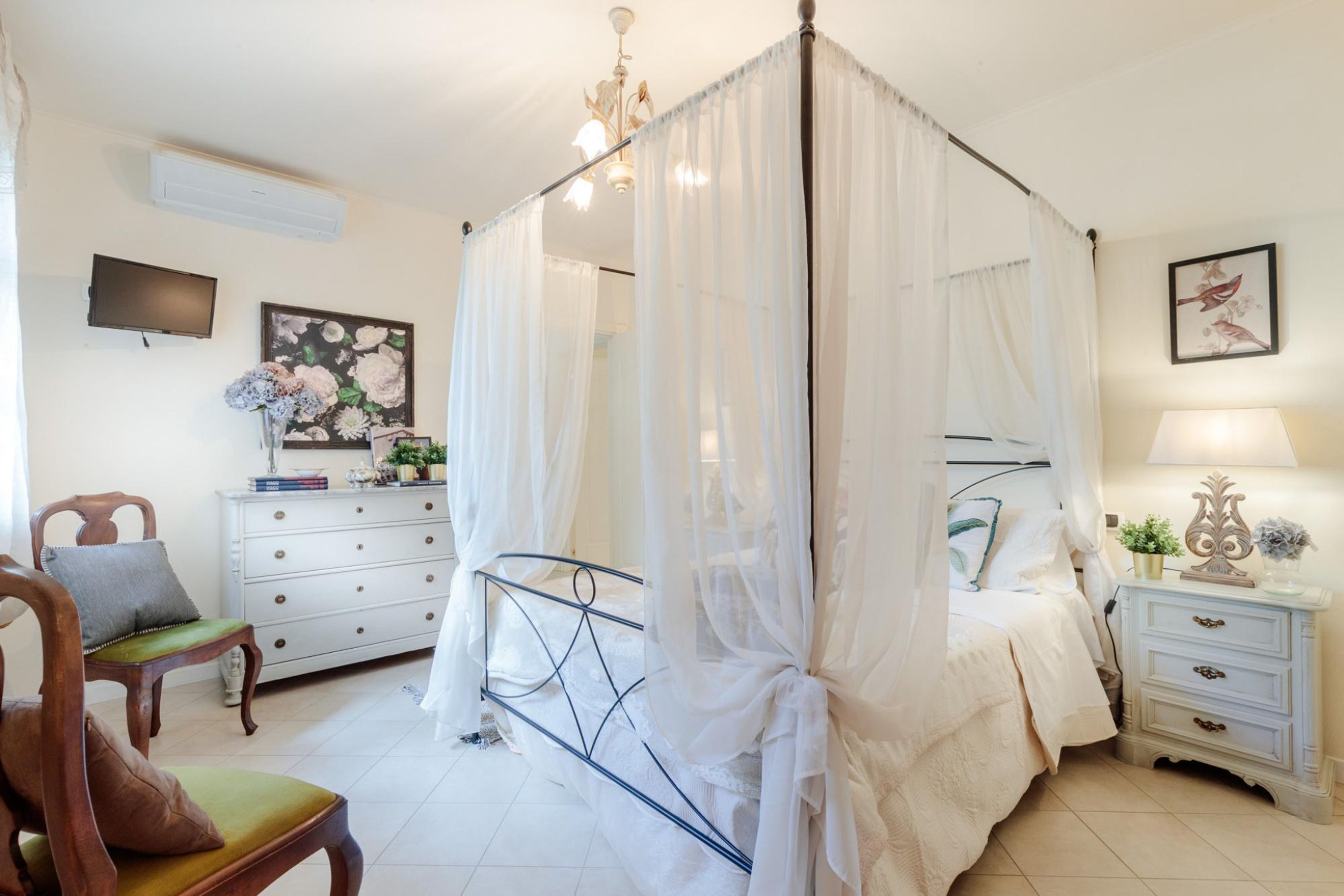 Een slaapkamer met een hemelbed, een nachtkastje met lamp, een kaptafel, een stoel en een televisie aan de muur.