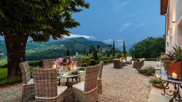 An outdoor dining area with wicker furniture, a table set with plates, glasses, and candles, surrounded by a gravel path and greenery. - Villa Regina in Lucca (Lucca)