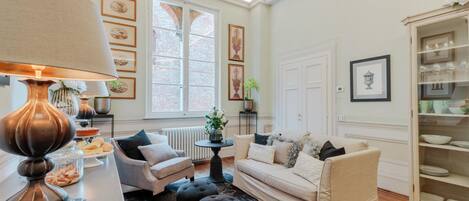 A well-lit living room with a sofa, armchairs, a coffee table, and a lamp. There are framed pictures on the walls and a window with a view.