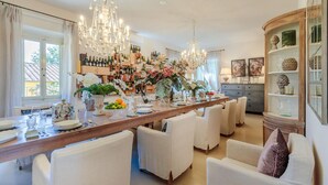 A spacious dining area with a long wooden table set for a meal, surrounded by white upholstered chairs. A chandelier hangs above, and a wine rack filled with bottles is visible in the background. - Villa Duemani - Pickleball Retreat (Capannori)