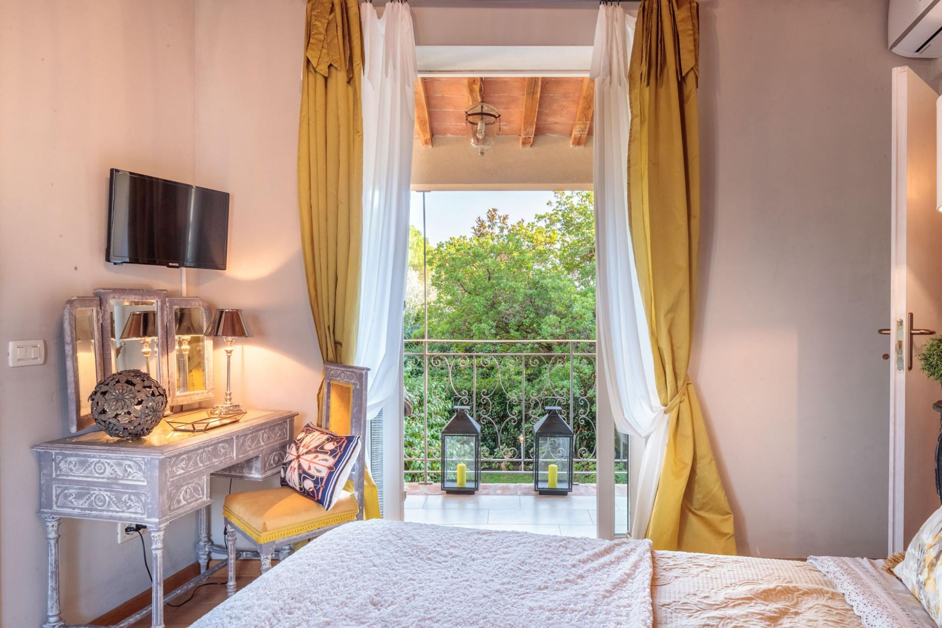 A bedroom with a bed, a bedside table, a TV mounted on the wall, and a balcony with yellow curtains.