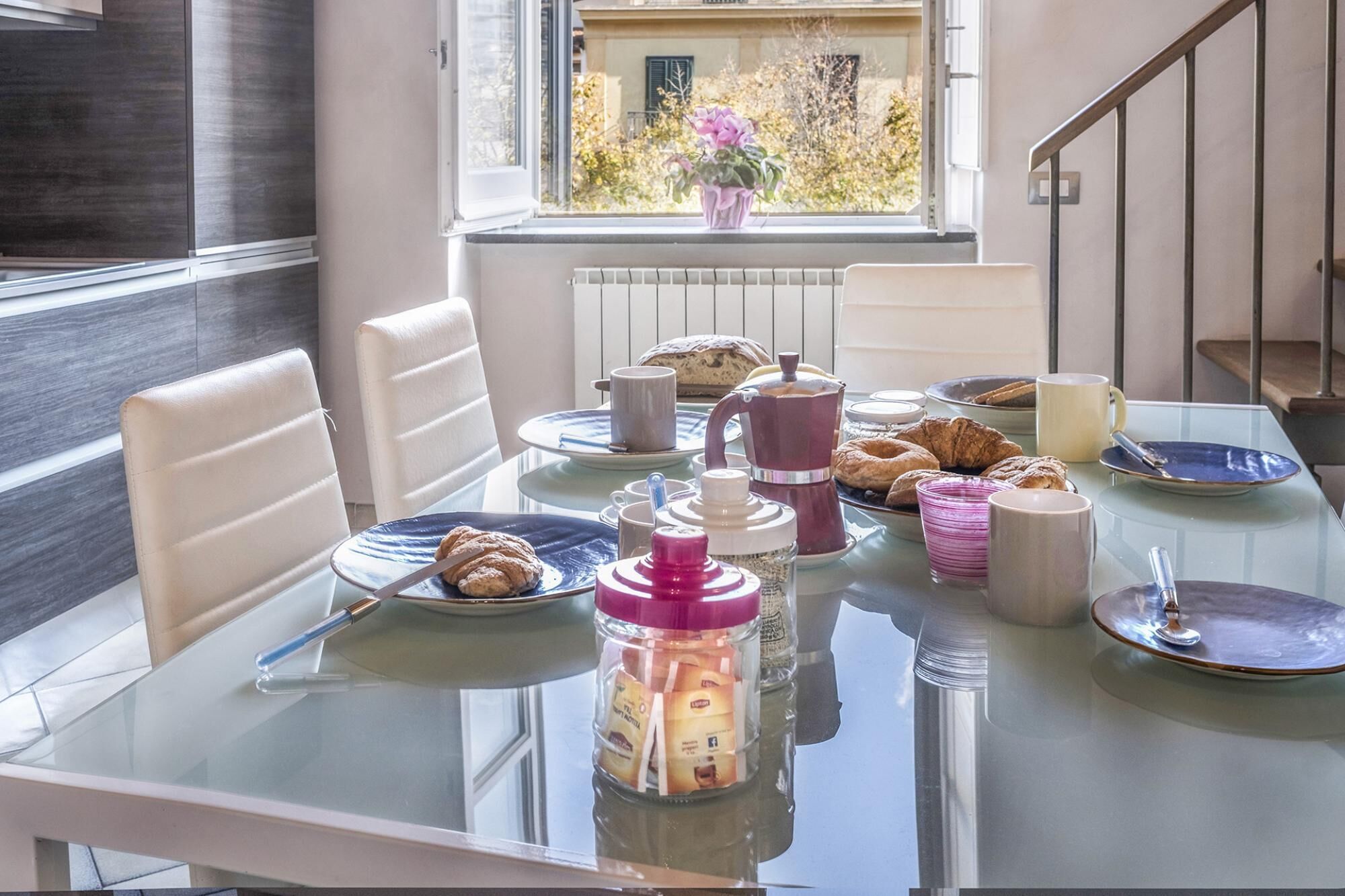 A breakfast table with pastries, coffee, and tea in a modern room with a view of a building and trees.