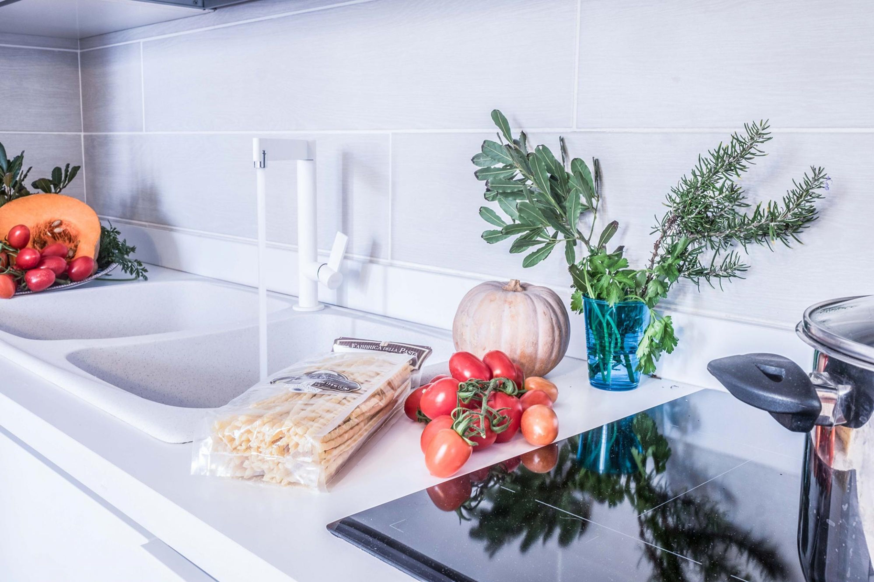 Eine Küchenarbeitsplatte mit Spüle, Schneidebrett, einer Packung Pasta, einer Kürbis, Tomaten und einer Topfpflanze.