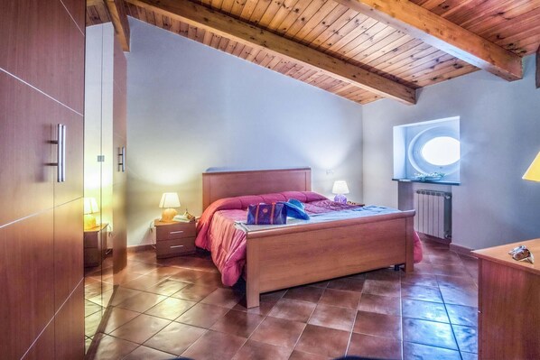 A bedroom with a wooden ceiling, a bed with pink bedding, a nightstand with a lamp, and a window with a circular view.