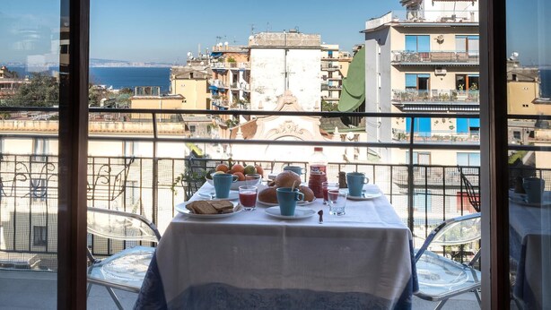 A balcony with a table set for breakfast, overlooking a cityscape with buildings and a view of the sea. - Appartamento Erica in Sorrento (Sorrento)