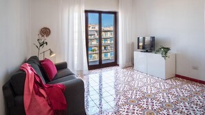 A living room with a sofa, a TV on a white cabinet, a patterned floor, and a view of buildings through a glass door. - Appartamento Erica in Sorrento (Sorrento)