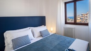 A bedroom with a bed, white pillows, a striped pillow, a blue bedspread, a nightstand with a lamp, and a window with a view of buildings and the sea. - Appartamento Erica in Sorrento (Sorrento)