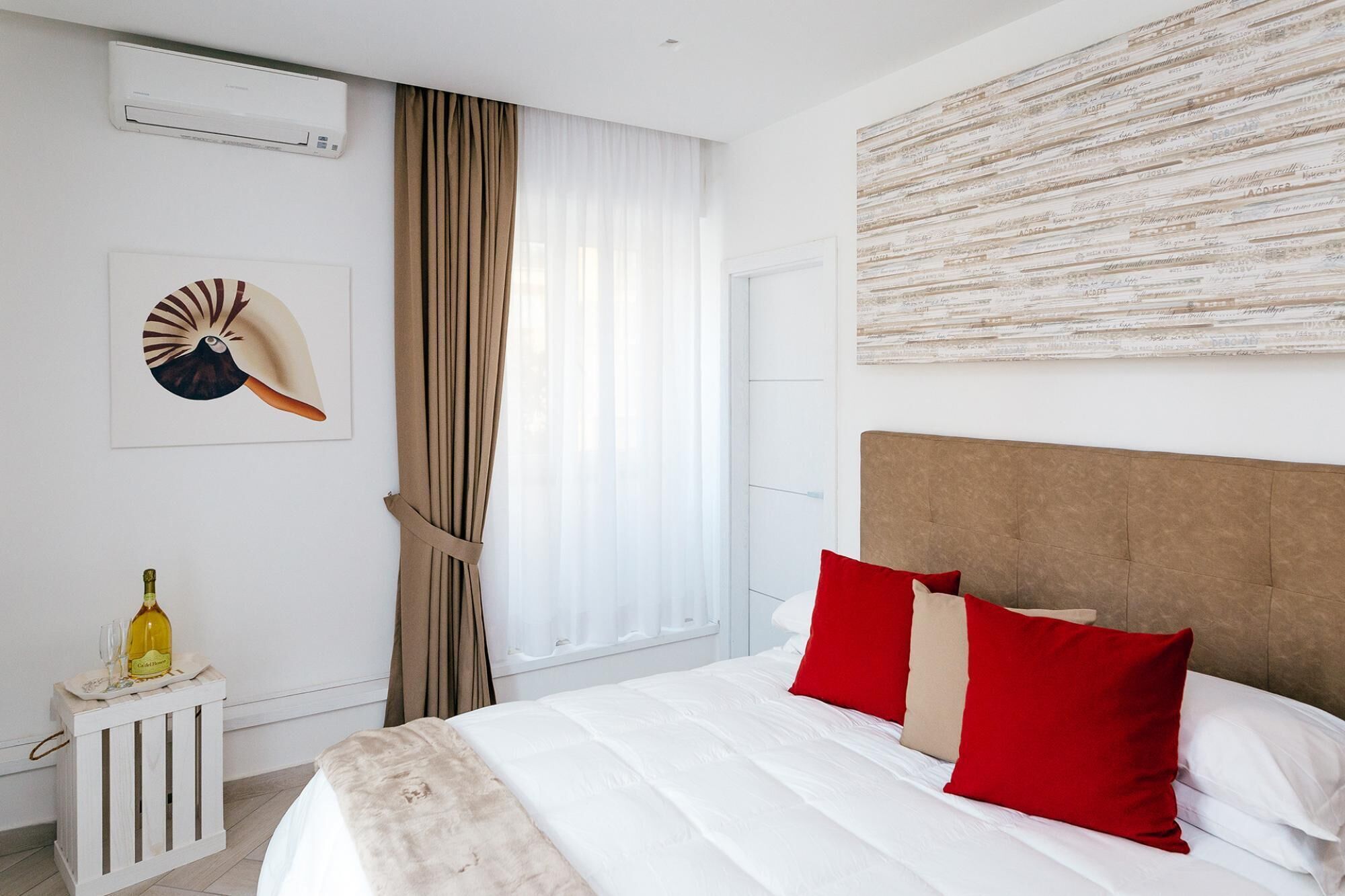 A hotel room with a large bed, a headboard, a wall-mounted air conditioner, a window with curtains, and a decorative piece on the wall.