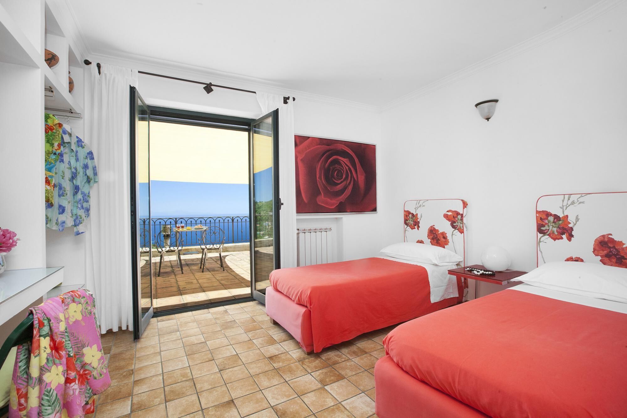 A hotel room with two beds, a balcony with a view, and a red rose painting.