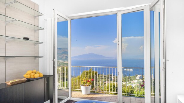 A modern room with a bed, a small kitchen area, and a balcony with a view of the sea and a mountain. - Casa Augusta A in Sorrento (Sorrento)
