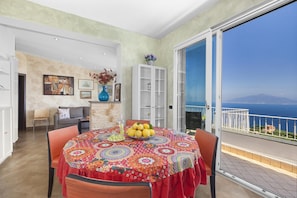 A dining area with a table set for four, a sofa, and a cabinet. A large window offers a view of the sea.