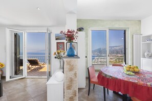 A dining area with a table set for two, a vase with flowers, and a view of the sea through large windows.