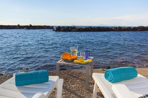 A beachside setting with two sun loungers, a table with a plate of fruit and drinks, and a view of the sea and rocky shore. - Villa del Sole in Massa Lubrense (Massa Lubrense)