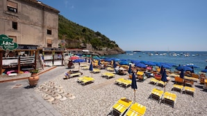 Un hôtel situé en bord de mer, doté d’une terrasse avec des tables et des parasols, d’une plage rocheuse et de bateaux amarrés dans l’eau.
