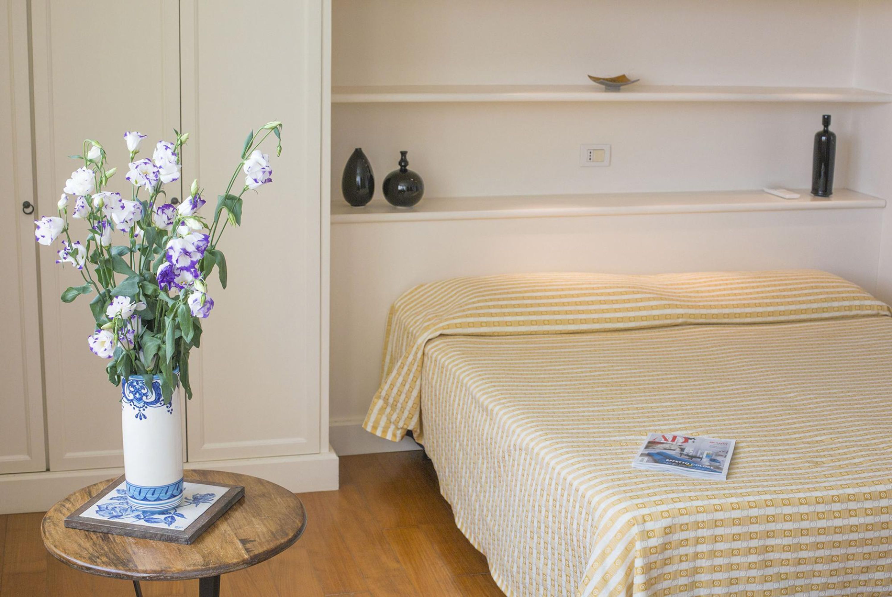 Una habitación con una cama, una mesita redonda con un florero y una repisa con adornos.