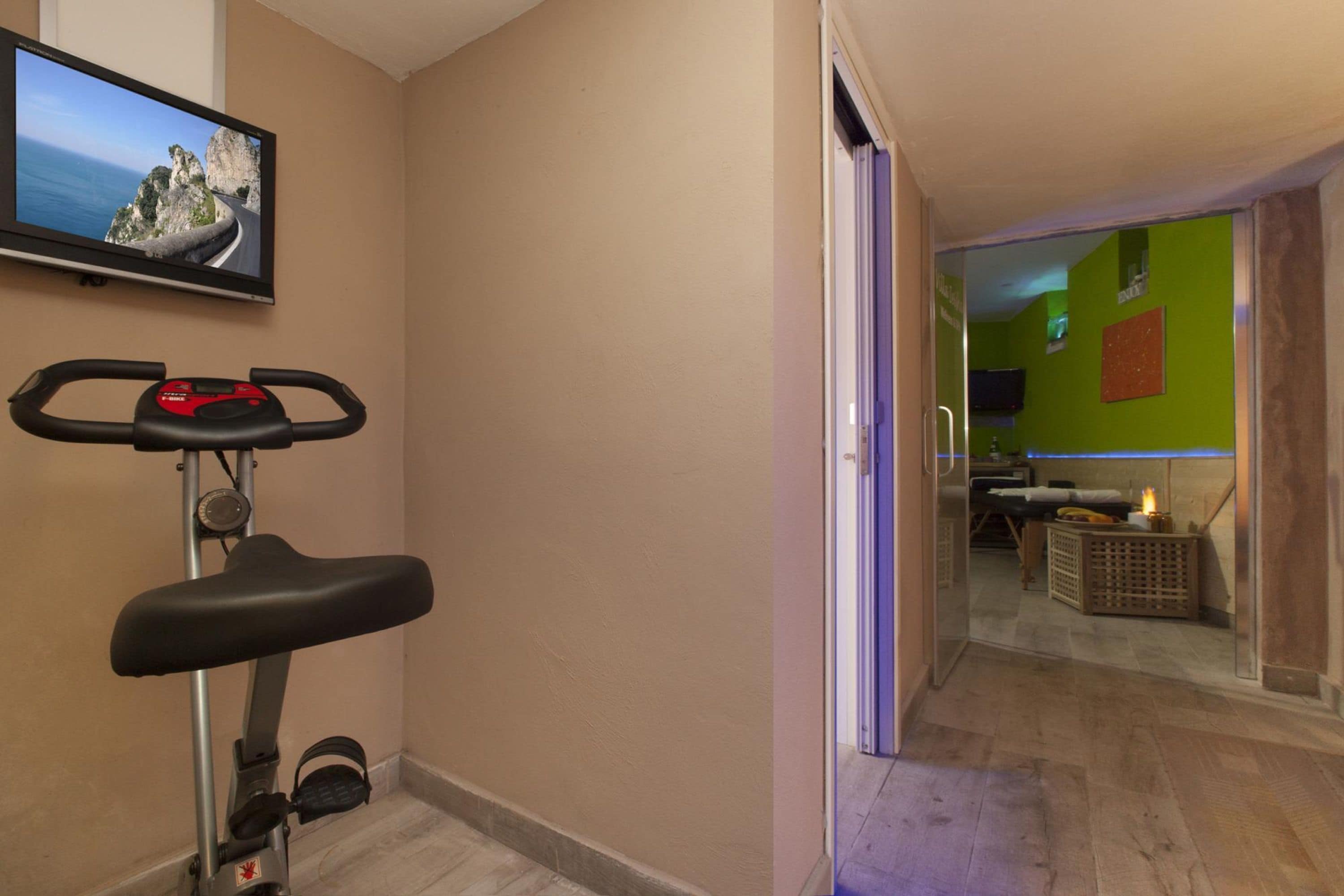 Quarto com bicicleta ergométrica, televisão exibindo uma paisagem e porta que dá para área iluminada em verde com uma cama e uma mesinha.