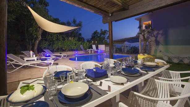 Uma área de refeições ao ar livre com piscina, cadeiras brancas e uma seção coberta com mesa posta para uma refeição.