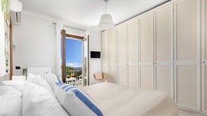 Quarto com cama, paredes brancas, guarda-roupa, uma cadeira e vista para o exterior.