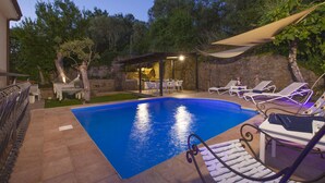Piscina com espreguiçadeiras e rede, cercada por um muro de pedra e vegetação.