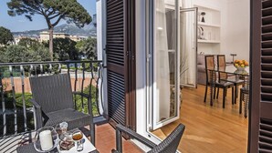 En balkon med fletningsmøbler, et bord med drikkevarer og udsigt til et træ og bygninger.