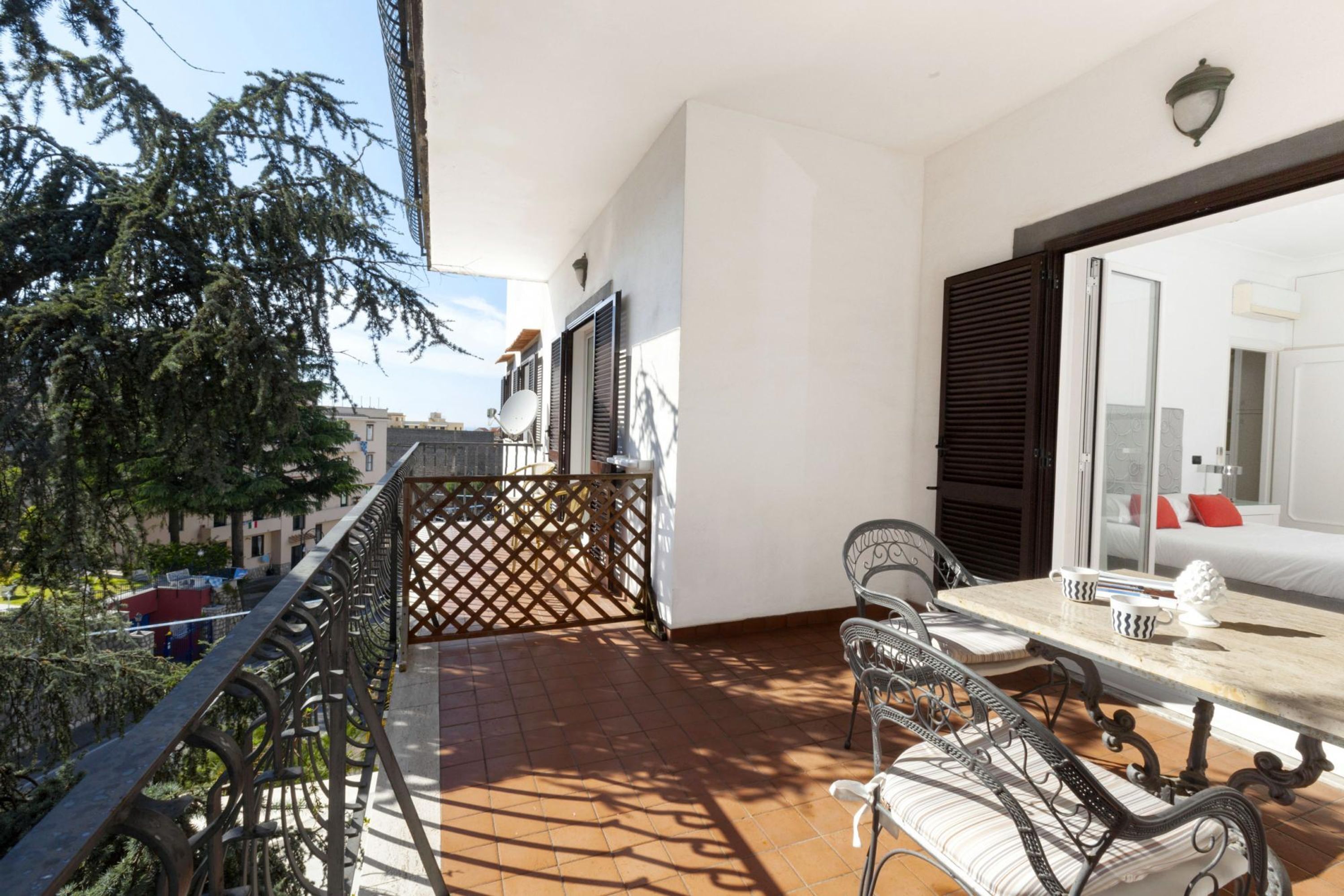 Un balcón con mesa y sillas, vistas a la ciudad y una puerta que da a una habitación con cama y ventana.