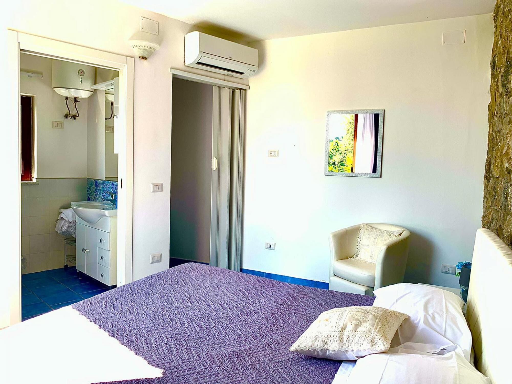 Quarto de hotel com cama, poltrona, banheiro com pia e ar condicionado.