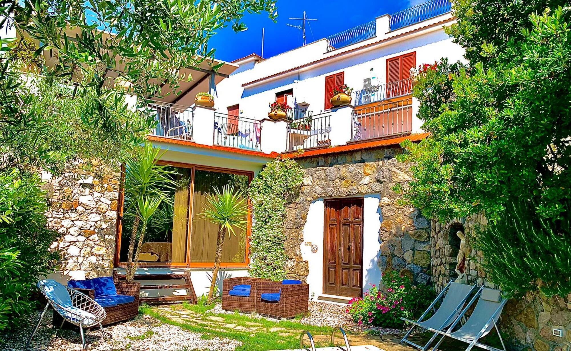Una casa de dos pisos con fachada de piedra, un balcón con contraventanas rojas y un jardín con mobiliario exterior.