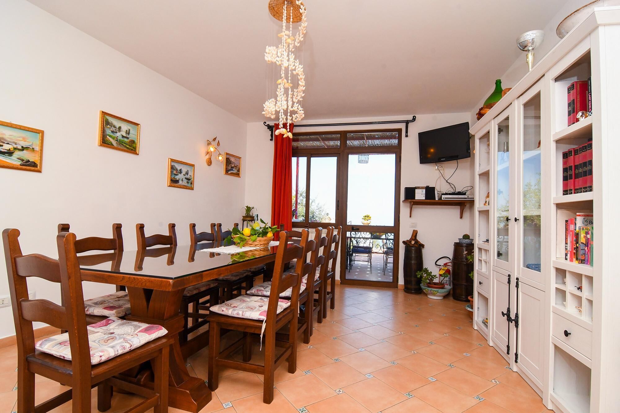 Uma área de refeições com mesa e cadeiras de madeira, televisão fixada na parede e estante repleta de livros.
