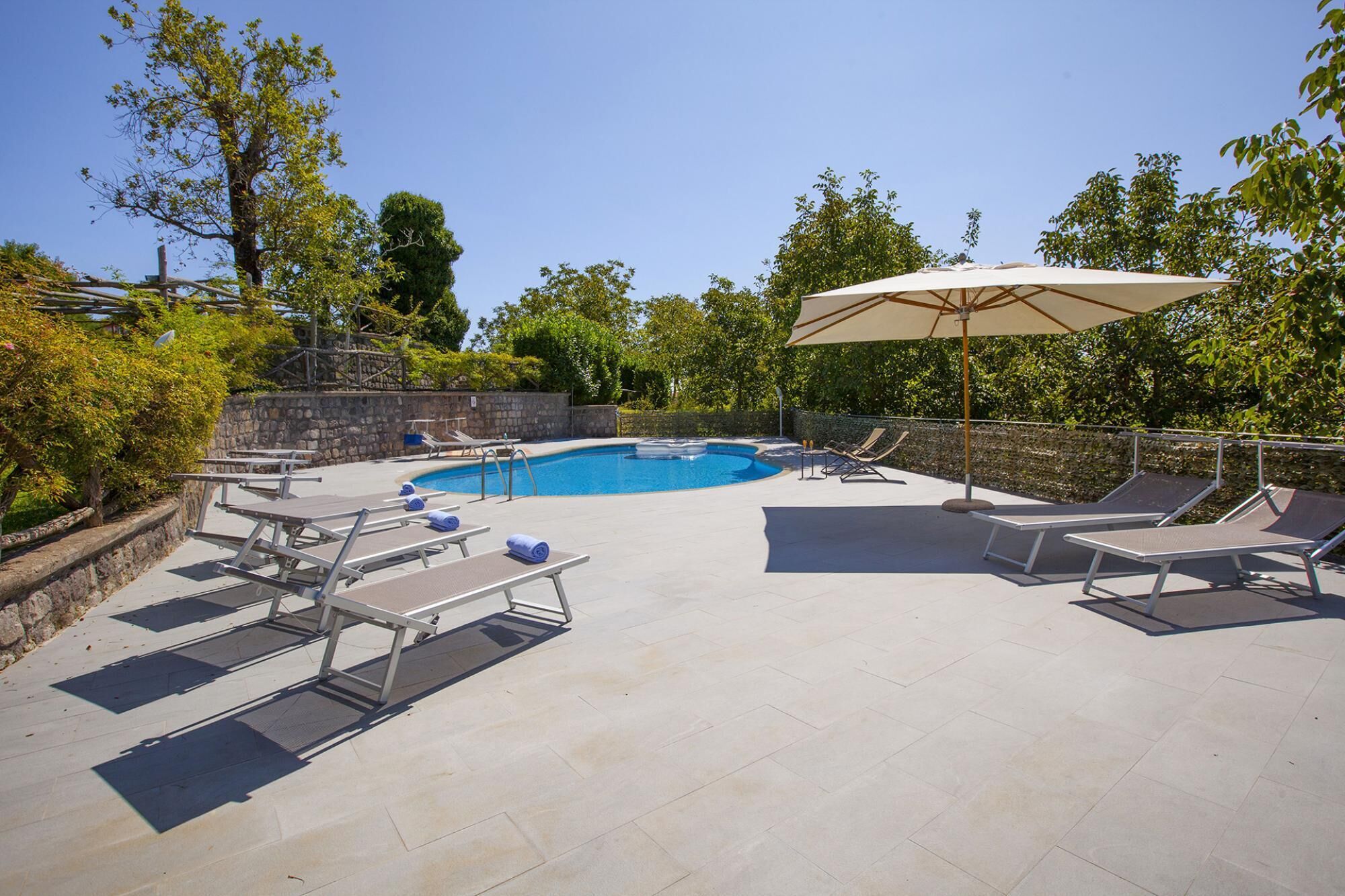 A swimming pool with lounge chairs and umbrellas, surrounded by stone walls and greenery.