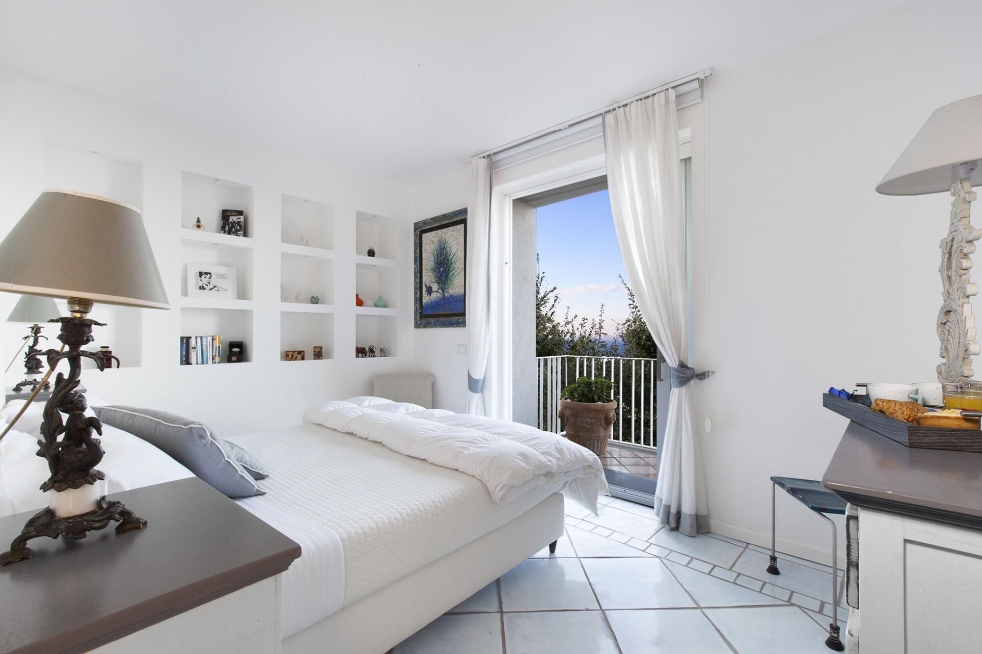 Un dormitorio con cama, mesita de noche, lámpara, estanterías y un balcón con vistas.